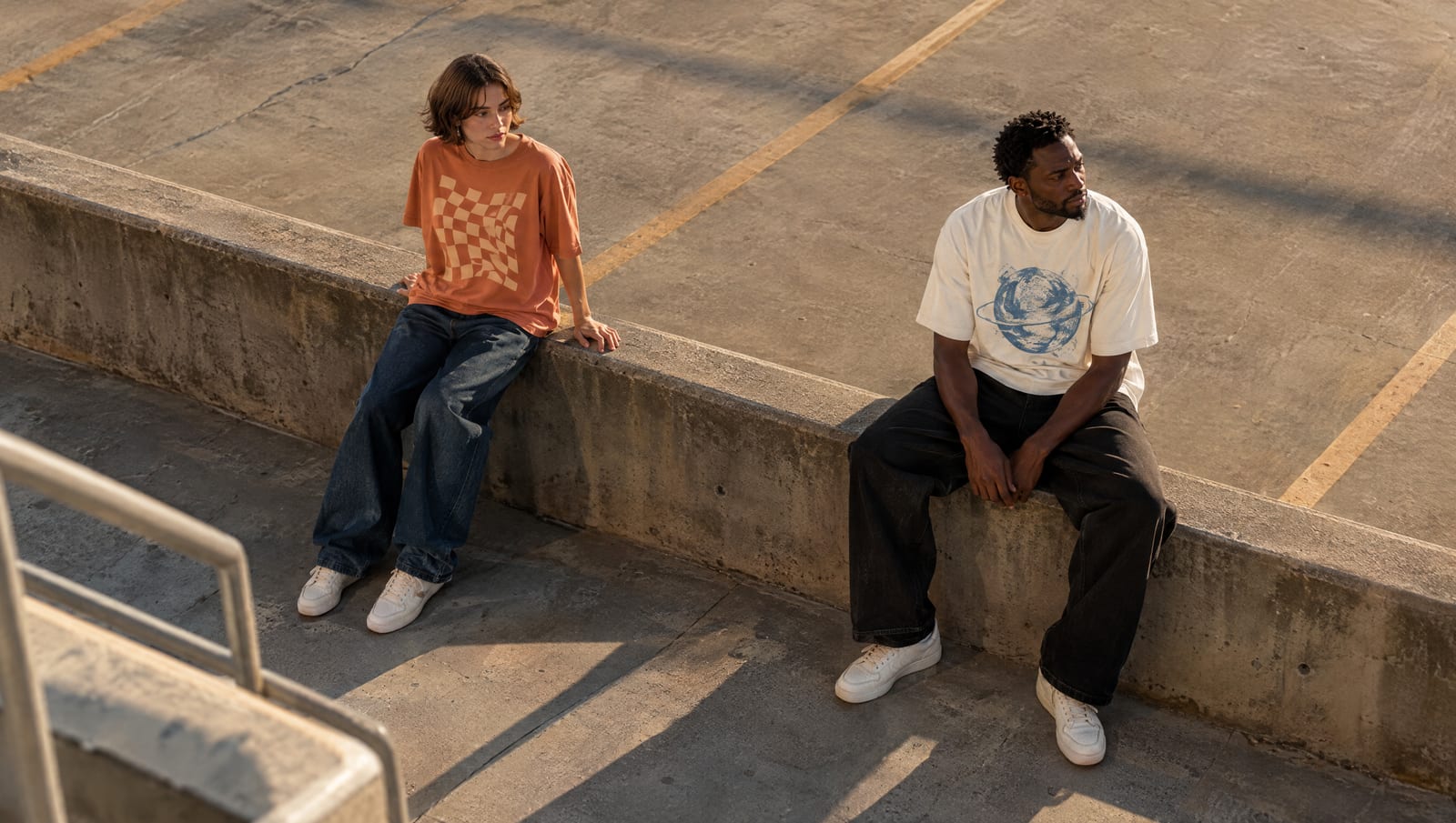 Two models sitting on a concrete ledge in a parking structure at golden hour, one in an orange checkerboard tee and baggy jeans, the other in a cream graphic tee and wide black trousers
