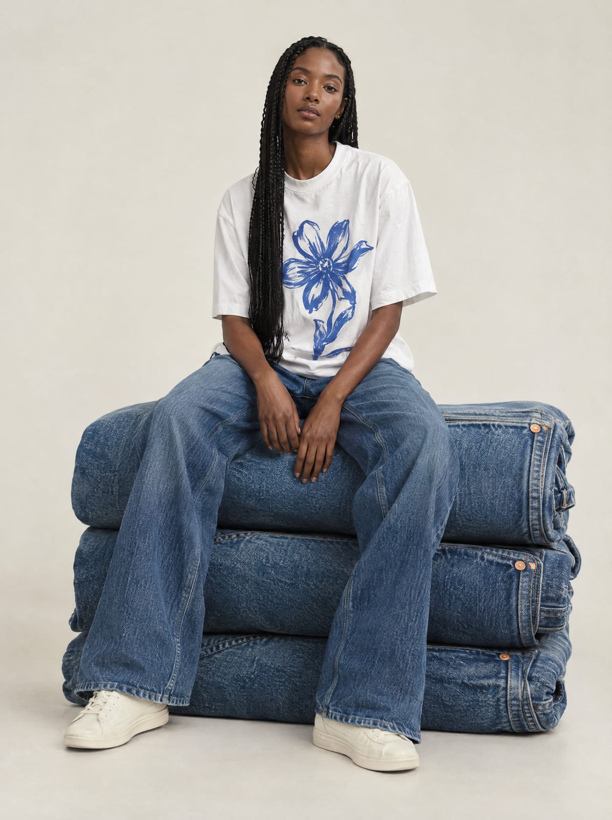 A model in a floral-print tee and wide jeans sitting on top of an oversized stack of folded denim
