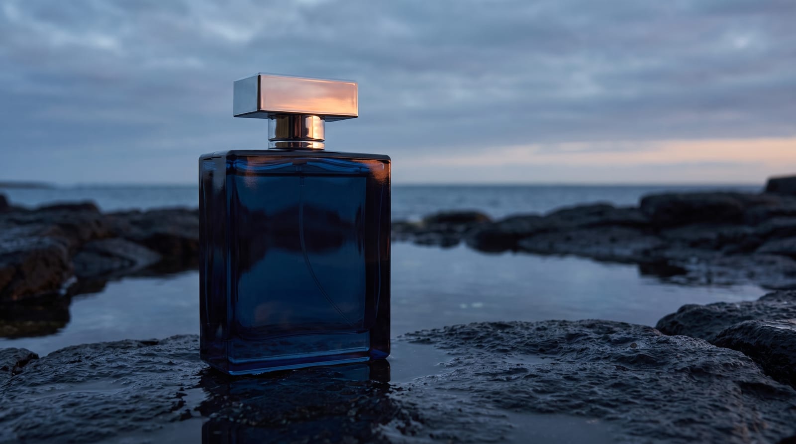 A deep blue glass fragrance bottle with a silver cap standing on wet rocks at dusk by the sea