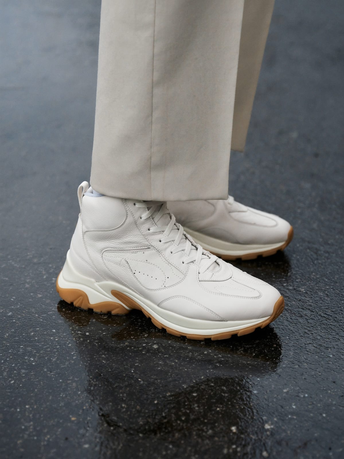 Cream leather high-top sneakers with gum soles on wet asphalt, cream trousers above